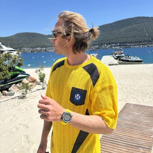 Puede incluir: Camiseta amarilla de manga corta con detalles negros y un escudo en el pecho. La camiseta tiene un patrón de punto texturizado. La persona lleva un reloj amarillo con esfera negra. El fondo incluye una playa, agua y montañas.