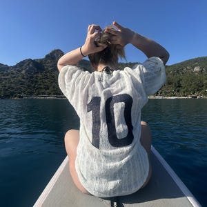 May include: A person wearing a white, open-weave shirt with the number 10 in black on the back, sitting on a paddleboard. The paddleboard is on the water, with mountains and a blue sky in the background.