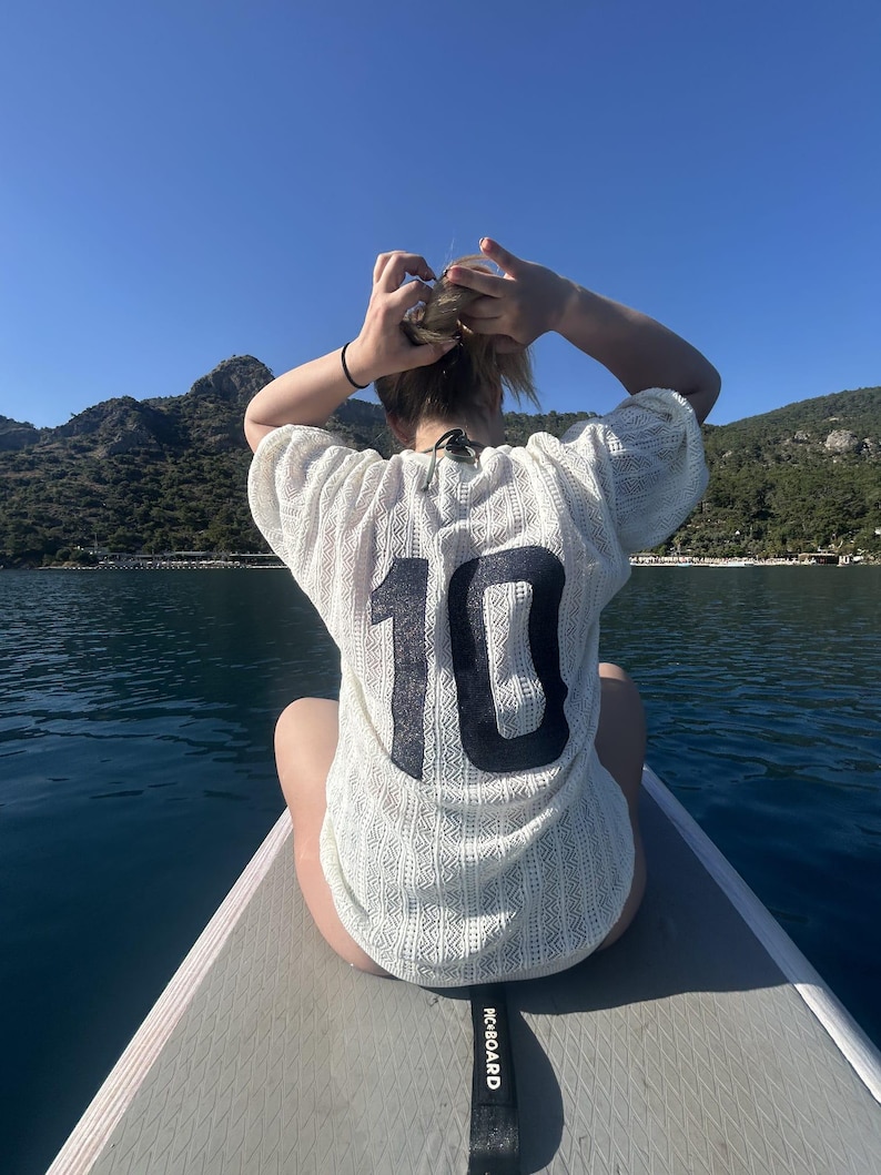 May include: A person wearing a white, open-weave shirt with the number 10 in black on the back, sitting on a paddleboard. The paddleboard is on the water, with mountains and a blue sky in the background.