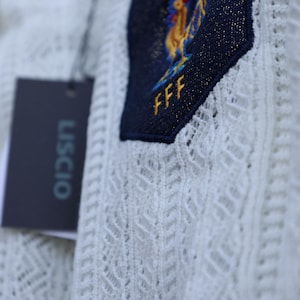 May include: Close-up of a white, open-weave fabric with a navy blue patch. The patch features a gold rooster emblem, two gold stars, and the letters "FFF". A black tag with the word "USCIO" is attached.