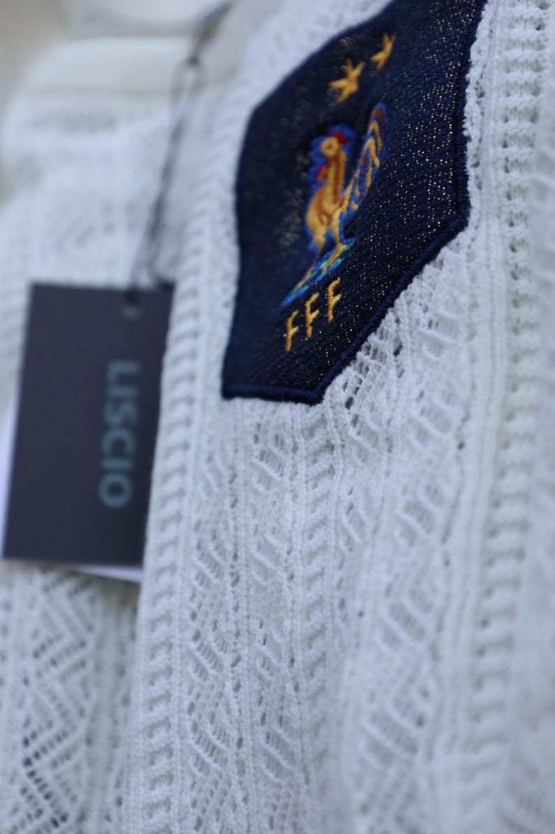 May include: Close-up of a white, open-weave fabric with a navy blue patch. The patch features a gold rooster emblem, two gold stars, and the letters "FFF". A black tag with the word "USCIO" is attached.