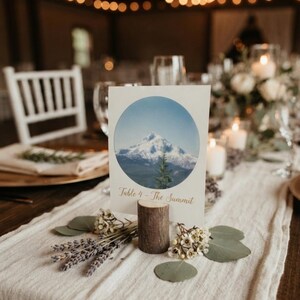 Peut inclure: Une carte de table rectangulaire blanche présentant une image circulaire d'une montagne enneigée et d'un sapin. Le texte "Table 4 - The Summit" est imprimé en écriture dorée sous le cercle. La carte est maintenue par un socle en bois cylindrique.