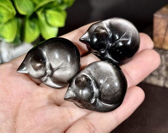 May include: Three small, polished obsidian carvings shaped like sleeping cats. Each figurine features a smooth, reflective surface with a metallic silver sheen, depicting a curled feline form with visible ears and a tucked tail.