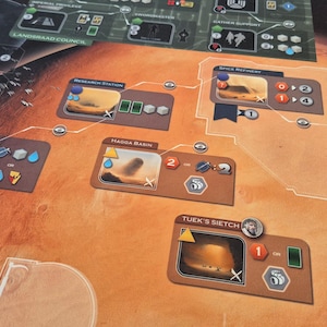 May include: A board game with a Martian landscape theme. The game board features various locations with illustrations and icons. Cards and tokens are scattered across the board, with a color palette of brown, orange, and green.