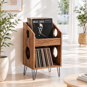 Puede incluir: Un mueble de almacenamiento de discos de madera con acabado marrón oscuro. La unidad tiene forma rectangular con recortes redondeados y patas de horquilla negras. La estantería superior muestra el álbum de Miles Davis, "Kind of Blue."