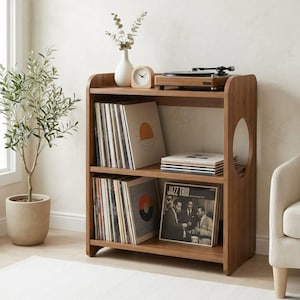 Puede incluir: Estantería de discos de madera con tres niveles, que contiene discos de vinilo. En la parte superior hay un tocadiscos, un reloj y un jarrón con vegetación. La estantería tiene un diseño redondeado y un recorte circular en el lateral.