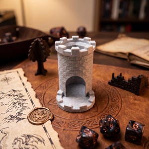 May include: A light grey, miniature castle tower dice holder with a crenellated top. The tower is made of stone-like material and sits on a brown leather-like surface with a map, wax seal, and several dice. A small tree and a broken wall are also visible.
