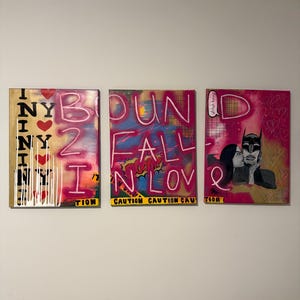 Op de afbeelding: Drie ingelijste kunstwerken met tekst en afbeeldingen in graffitistijl. Het linkerpaneel zegt "I NY" met harten en druppels. Het middelste paneel luidt "BOUND FALL IN LOVE" met een stripboekachtige "BOOM"-bel. Het rechterpaneel toont een Batman-achtig figuur.