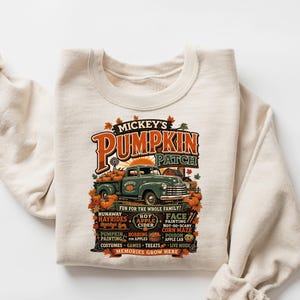 Könnte beinhalten: Ein cremefarbenes Sweatshirt mit Rundhalsausschnitt und einem Grafikdruck eines grünen Oldtimer-Pickups, der mit Kürbissen beladen ist. Der Text lautet Mickey's Pumpkin Patch, Fun for the whole family und listet herbstliche Aktivitäten.