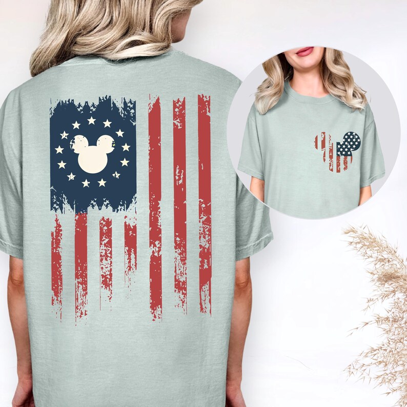 May include: A light sage green t-shirt featuring a large distressed American flag graphic on the back with a central Mickey Mouse silhouette surrounded by stars. The front chest area displays a smaller Mickey Mouse head filled with a flag pattern.