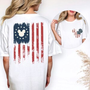 May include: A white t-shirt featuring a distressed American flag graphic on the back with a central mouse head silhouette surrounded by stars. The front chest area displays a smaller, matching mouse head graphic filled with a red, white, and blue flag pattern.