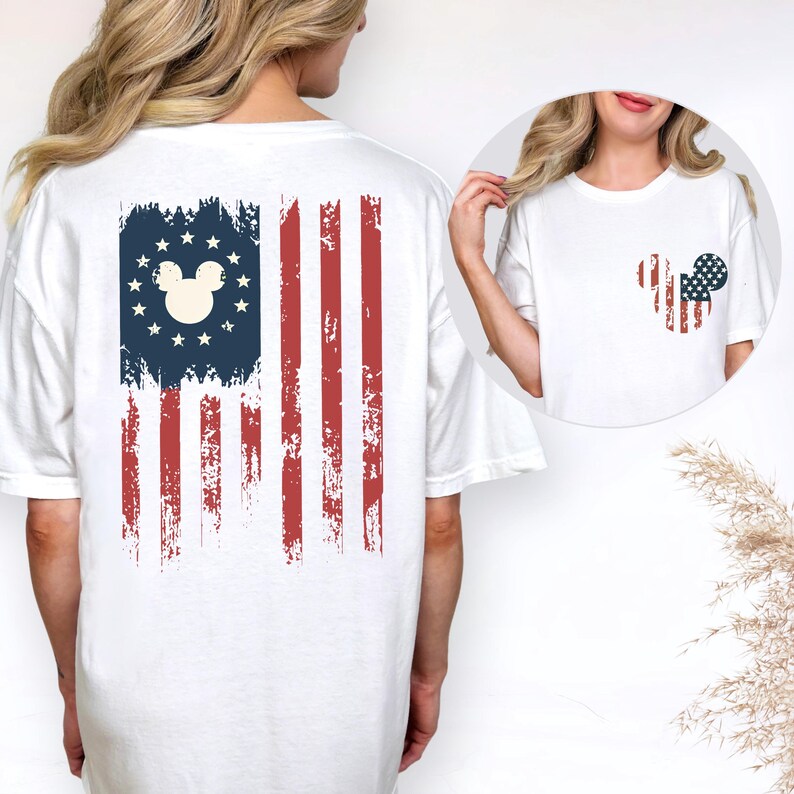 May include: A white t-shirt featuring a distressed American flag graphic on the back with a central mouse head silhouette surrounded by stars. The front chest area displays a smaller, matching mouse head graphic filled with a red, white, and blue flag pattern.