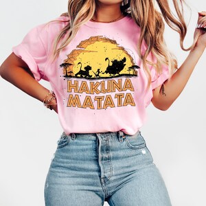 Puede incluir: Una camiseta de manga corta de color rosa claro con un estampado gr&aacute;fico de tres animales de dibujos animados caminando por un paisaje frente a un gran sol amarillo. El texto HAKUNA MATATA aparece en letras naranjas.