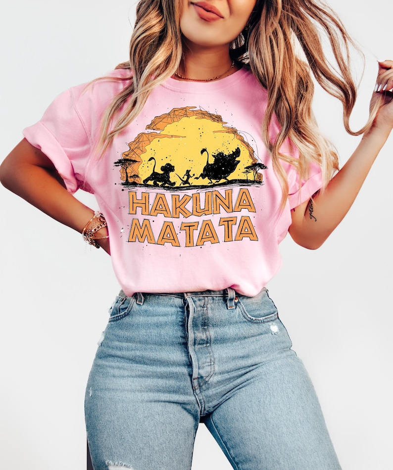Puede incluir: Una camiseta de manga corta de color rosa claro con un estampado gr&aacute;fico de tres animales de dibujos animados caminando por un paisaje frente a un gran sol amarillo. El texto HAKUNA MATATA aparece en letras naranjas.
