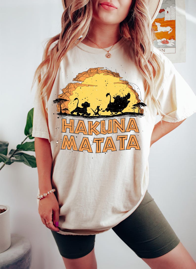 Puede incluir: Una camiseta de manga corta color crema con un estampado gr&aacute;fico de tres siluetas caminando por un paisaje frente a un gran sol amarillo. El texto "HAKUNA MATATA" est&aacute; impreso en letras naranjas llamativas debajo del gr&aacute;fico con &aacute;rboles.