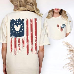 May include: A cream-colored t-shirt featuring a large graphic on the back of a distressed American flag with a white Mickey Mouse silhouette centered in the blue field of stars. A smaller version of the same flag-patterned mouse icon is on the front.