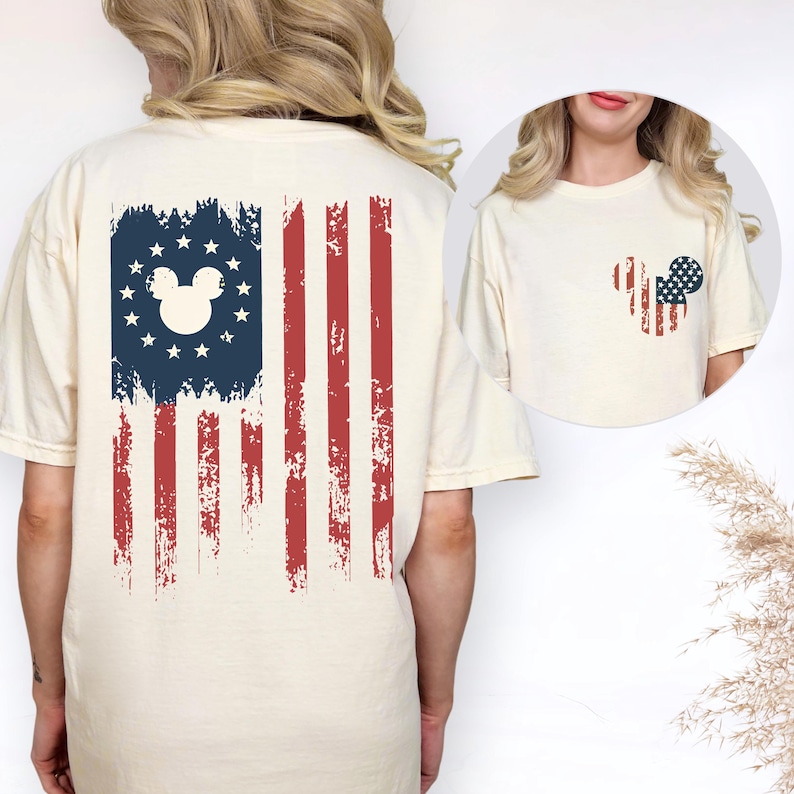 May include: A cream-colored t-shirt featuring a large graphic on the back of a distressed American flag with a white Mickey Mouse silhouette centered in the blue field of stars. A smaller version of the same flag-patterned mouse icon is on the front.