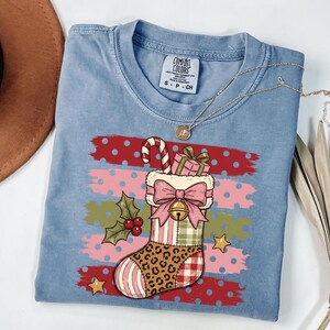 May include: A blue short-sleeve t-shirt with a graphic print of a Christmas stocking featuring a leopard print toe, a pink bow, a candy cane, and a gift. The background of the graphic has horizontal brush strokes in red, pink, and green with polka dots.