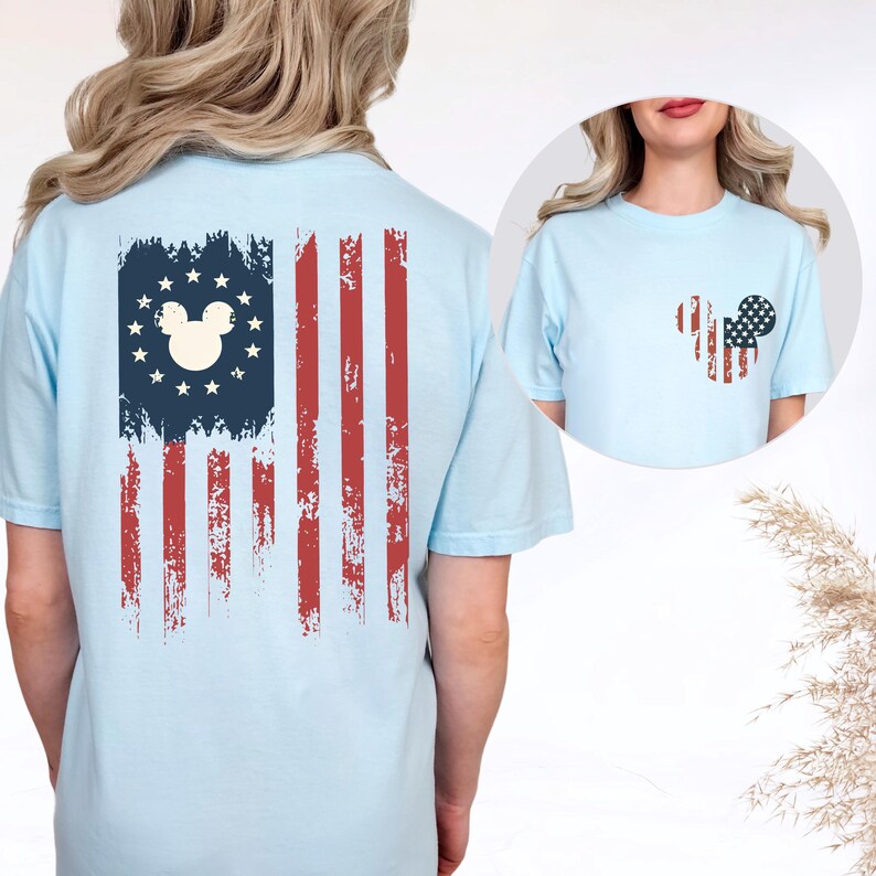 May include: A light blue t-shirt featuring a distressed American flag graphic on the back with a central mouse head silhouette surrounded by stars. The front chest area displays a smaller, matching mouse head graphic with a flag pattern.