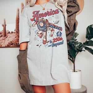 May include: A light grey t-shirt featuring a graphic of a space ranger character in a red, white, and blue suit. The text reads America, To 250th and Beyond, with star and planet illustrations. The shirt has short sleeves and a crew neckline.
