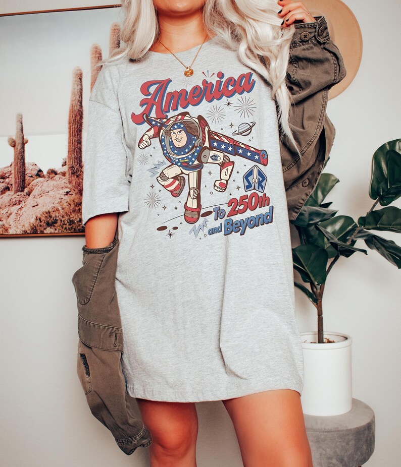 May include: A light grey t-shirt featuring a graphic of a space ranger character in a red, white, and blue suit. The text reads America, To 250th and Beyond, with star and planet illustrations. The shirt has short sleeves and a crew neckline.