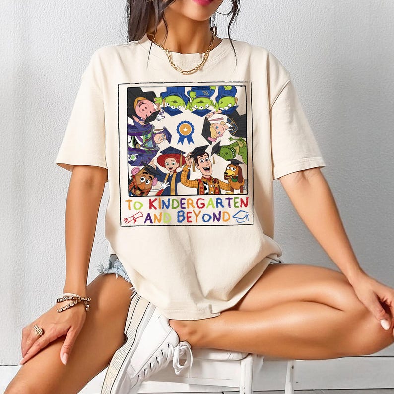 Pu&ograve; includere: Una t-shirt beige a maniche corte con una stampa grafica quadrata di vari personaggi dei cartoni animati che indossano il tocco da laurea. Il testo sotto i personaggi recita "TO KINDERGARTEN AND BEYOND" in lettere colorate, con icone di diploma e tocco.