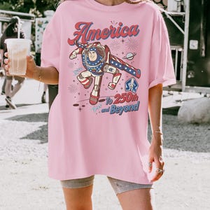 May include: A light pink t-shirt featuring a graphic of a space ranger character in a red, white, and blue suit. The text reads America To 250th and Beyond, accompanied by illustrations of fireworks, a planet, and stars in a patriotic colour scheme.