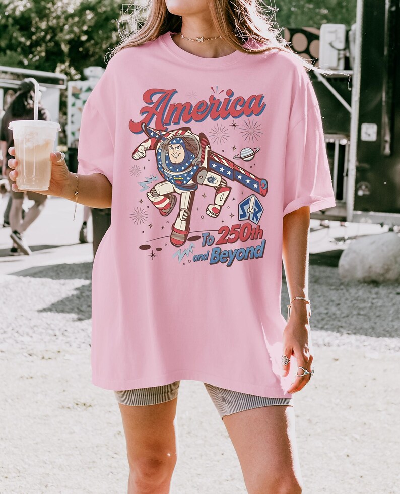 May include: A light pink t-shirt featuring a graphic of a space ranger character in a red, white, and blue suit. The text reads America To 250th and Beyond, accompanied by illustrations of fireworks, a planet, and stars in a patriotic colour scheme.