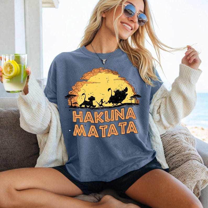 Puede incluir: Una camiseta azul pizarra con un estampado gr&aacute;fico de una silueta de sol amarillo con contornos negros de un cachorro de le&oacute;n, un suricato y un jabal&iacute; en una sabana, con el texto naranja HAKUNA MATATA impreso debajo del gr&aacute;fico.