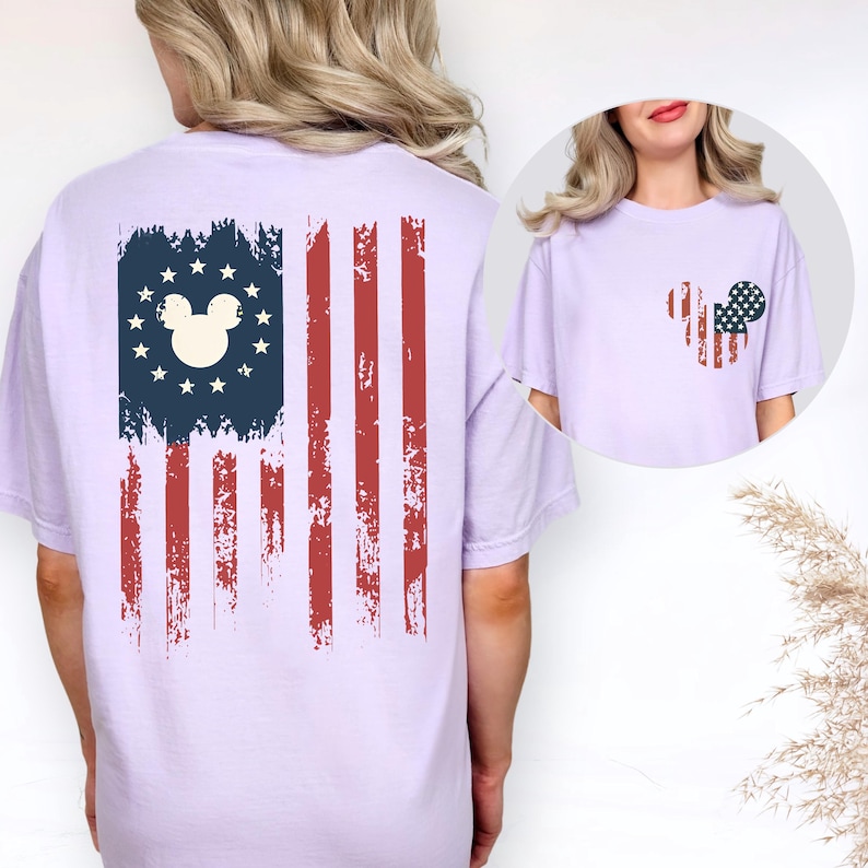 May include: A light purple short-sleeve t-shirt featuring a distressed American flag graphic on the back with a central Mickey Mouse silhouette. The front chest area displays a smaller, matching distressed flag graphic in a Mickey Mouse shape.