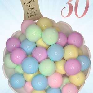 May include: A white scalloped bowl filled with thirty smooth, spherical plastic capsules in pastel shades of pink, blue, yellow, and green. A small wooden easel stands behind the bowl, displaying a card with the text, If You Don't Try, You Never Know.