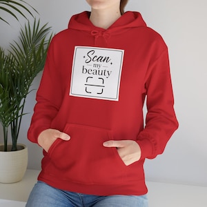 Puede incluir: Una sudadera con capucha roja con un bolsillo delantero. La sudadera presenta un gráfico cuadrado blanco con el texto "Scan my beauty" y un símbolo de código de barras en negro. La persona lleva vaqueros azules.