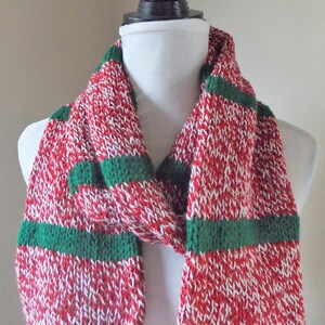 May include: A knitted scarf with a red and white speckled pattern, accented with green stripes. The scarf is displayed on a white mannequin head. The scarf is made of a soft, textured material.