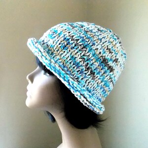 May include: A knitted cloche hat in shades of blue, white, and brown. The hat features a small brim and a textured knit pattern. This handmade hat is a stylish accessory for cooler weather.