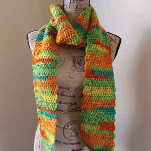 May include: A handmade, colorful crochet scarf displayed on a mannequin. The scarf features a vibrant mix of orange, yellow, green, and teal stripes. The scarf is made of soft yarn and has a textured appearance, perfect for adding a pop of color to any outfit.