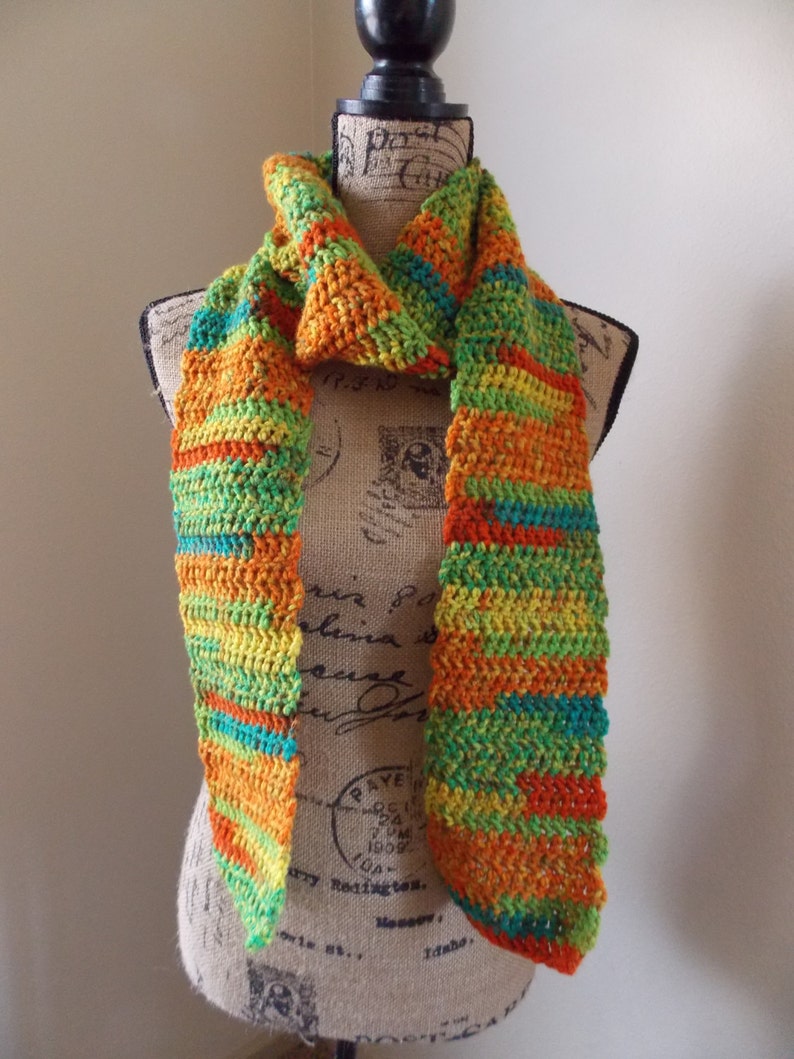 May include: A handmade, colorful crochet scarf displayed on a mannequin. The scarf features a vibrant mix of orange, yellow, green, and teal stripes. The scarf is made of soft yarn and has a textured appearance, perfect for adding a pop of color to any outfit.
