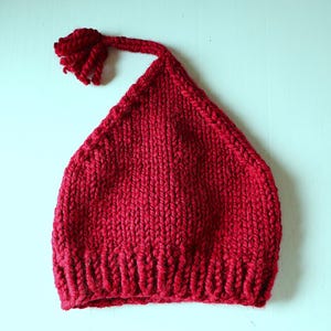 May include: A knitted red hat with a pointed top and a tassel. The hat is made of a thick, textured yarn and has a ribbed brim. The hat is a solid, deep red color and is likely handmade.