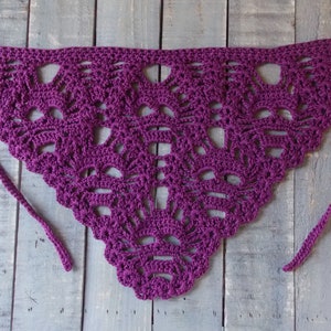 May include: A handmade, purple crochet shawl with a triangular shape and intricate openwork design. The shawl features a scalloped edge and long ties for securing. The openwork pattern creates a delicate, airy look. The shawl is displayed on a wooden surface.