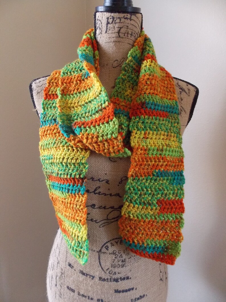 May include: A handmade crochet scarf in a vibrant mix of colors, including orange, yellow, green, and teal. The scarf is displayed on a mannequin, showcasing its textured, striped pattern. This accessory is perfect for adding a pop of color to any outfit.