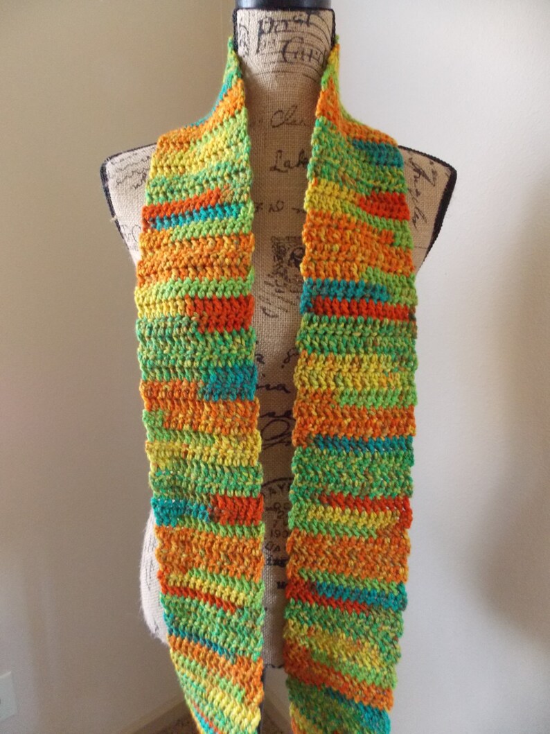 May include: A colorful, handmade crochet scarf. The scarf features horizontal stripes of orange, yellow, green, and teal. The scarf is displayed on a mannequin. This accessory is perfect for adding a pop of color to any outfit.