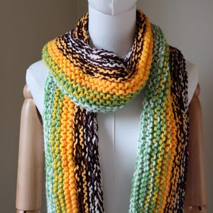 May include: A hand-knitted scarf featuring a chunky garter stitch pattern with vertical stripes in vibrant shades of yellow, light green, dark brown, and cream, draped around a white mannequin neck and shoulders.