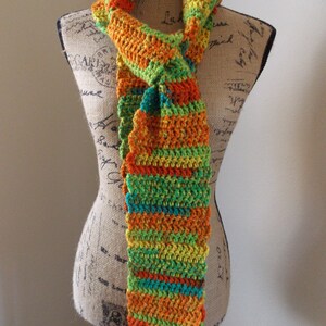 May include: A colorful, handmade crochet scarf in shades of orange, green, yellow, and teal. The scarf is displayed on a mannequin and features a textured, striped pattern. The scarf is a unique accessory.