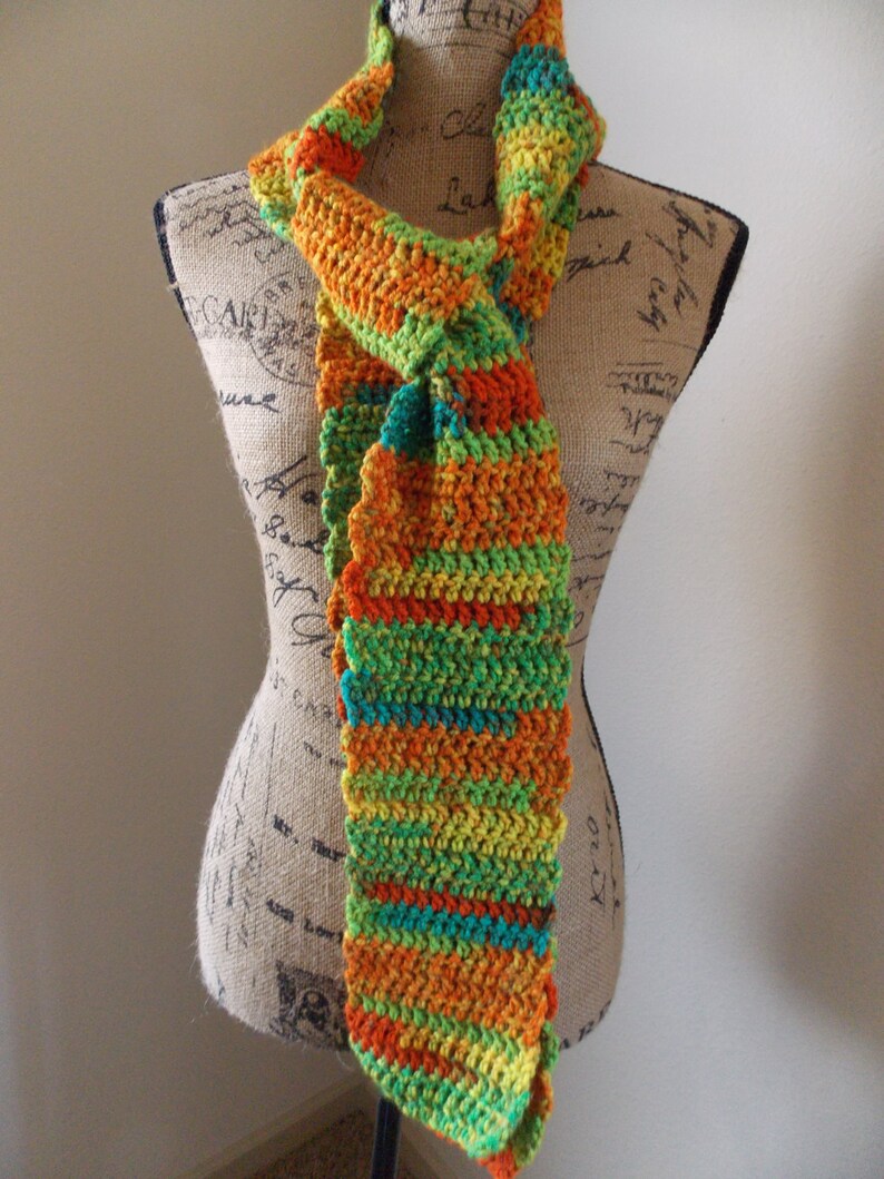 May include: A colorful, handmade crochet scarf in shades of orange, green, yellow, and teal. The scarf is displayed on a mannequin and features a textured, striped pattern. The scarf is a unique accessory.