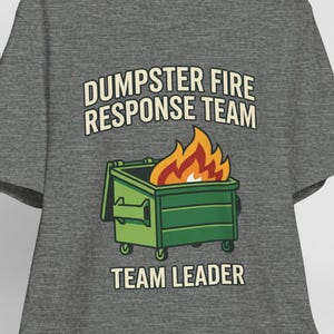 May include: A heather gray short-sleeve t-shirt featuring a graphic of a green dumpster with orange and yellow flames. The text reads DUMPSTER FIRE RESPONSE TEAM above the dumpster and TEAM LEADER below it. A white baseball cap hangs nearby.