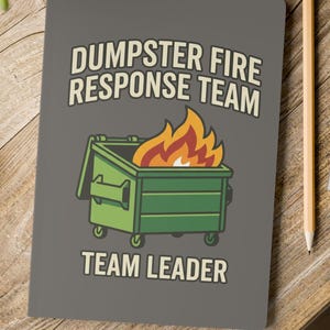 May include: A gray notebook featuring a green dumpster graphic with orange and yellow flames emerging from the top. The text DUMPSTER FIRE RESPONSE TEAM is printed above the dumpster, and the words TEAM LEADER are printed below in light lettering.