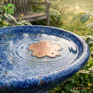 May include: A round, blue and white speckled ceramic bird bath filled with water. A flat, copper-colored metal flower ornament with five petals and a central stamen design floats on the surface of the water, creating subtle concentric ripples.