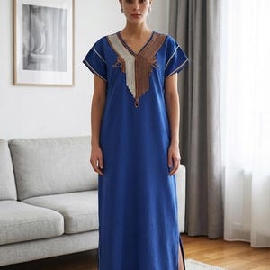 Könnte beinhalten: Ein bodenlanges blaues Kaftankleid mit kurzen Ärmeln, einem V-Ausschnitt und einem dekorativen Brustbereich mit kontrastierenden beigen und braunen strukturierten Stickereien. Das Kleidungsstück ist mit weißen Paspeln an Ärmeln und Saum versehen.