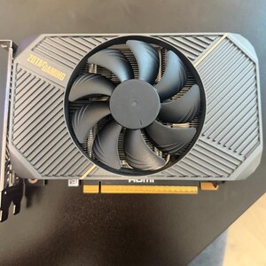 May include: A ZOTAC GAMING graphics card with a black fan and a gray metal casing. The card has a gold-colored connector and the text "ZOTAC GAMING" is visible on the top left.