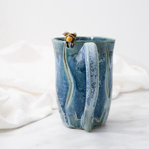 May include: A handmade ceramic mug with a mottled blue glaze and vertical ridges. A small, three-dimensional yellow and black bee figure is attached to the top rim of the mug, positioned directly above the curved handle on the side of the vessel.
