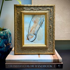 May include: A framed artwork with a gold ornate frame. The artwork features a sketch with black and brown tones. The frame sits atop two books, one with the text "RH INTERIORS SPRING 2026" and the other "OIL PAINTER'S COLOR HANDBOOK".